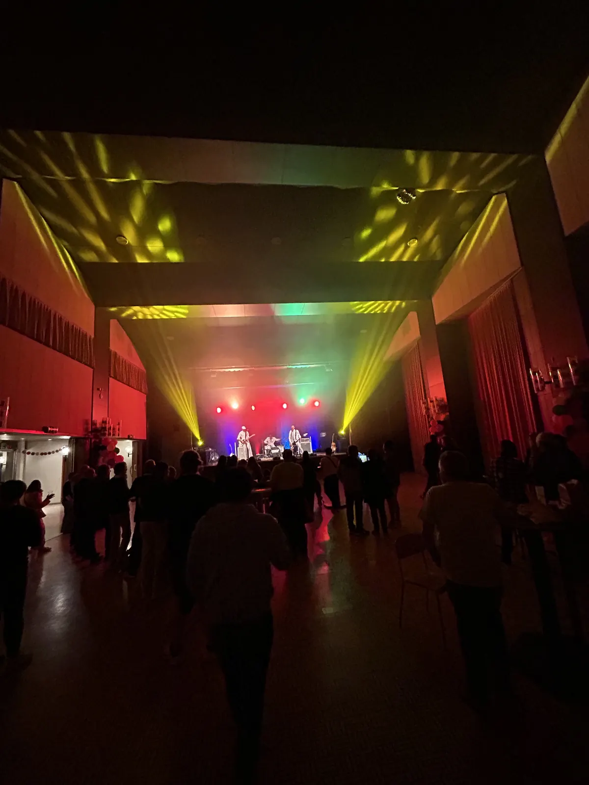 Concert sur la scène du Centre communal de Péry, éclairée en rouge, vert et bleu, avec des faisceaux de lyres jaunes projetés au plafond au-dessus du public